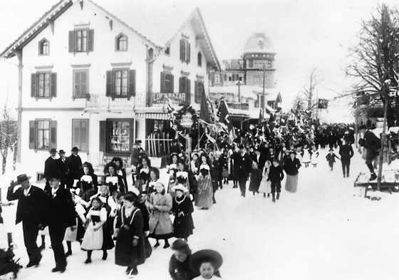 Das Schweizerische Skirennen von 1910 mit dem obligaten Festumzug. Eine lockere Parade mit schönen Trachten, vielen Kindern und würdigen Gemeinderäten. Noch lange sprach man davon. Im Hintergrund Luf’s Bazar von Peter Bernet, wo Ski auf Käufer warten.